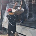 Zwei Personen laden einen großen Sack aus einem Lastwagen, der mit gestapelten Säcken beladen ist. Eine Person steht im Lastwagen und reicht den Sack einer anderen Person draußen. Ein Hund steht in der Nähe und beobachtet das Geschehen.