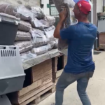 Ein Mann in einem blauen T-Shirt und einer roten Baseballkappe hebt eine schwere Plastiktüte mit Kies von einer gestapelten Palette.