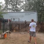 Ein Mann in einem weißen T-Shirt geht auf einen eingezäunten Bereich zu, im Hintergrund steht ein großer, geparkter Anhänger.