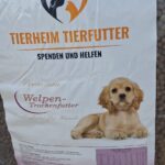 Ein großer Sack Hundefutter der Marke „Tierheim Tierfutter“ mit dem Foto eines Welpen, Spendenaufrufen und Hilfsaufforderungen auf Deutsch sowie sichtbaren braunen Trockenfutterbrocken im Inneren des Sacks.