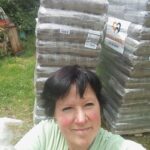 Eine Frau mit kurzen, dunklen Haaren lächelt draußen neben zwei großen, verpackten Warenpaletten. Links ist ein Teil des Schwanzes eines weißen Hundes zu sehen. Im Hintergrund sind Gras und gestapelte Gegenstände zu erkennen.