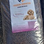Ein großer Sack mit „Tierheim Tierfutter“-Hundefutter, auf dessen Etikett ein goldfarbener, liegender Welpe und deutscher Text abgebildet sind. Der Sack ist mit braunen Trockenfutterbrocken gefüllt und steht auf Kies.
